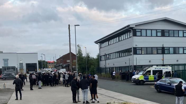 Crowd of people in Oldham with a police van over the road