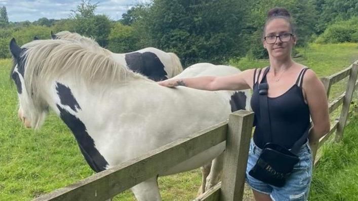 Kelly Reilly wearing a black vest with denim shorts standing next to a fence with her arm outstretched touching a white horse with black spots