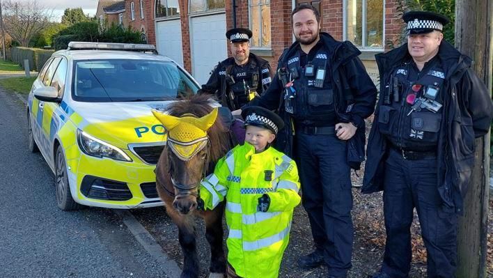 Three police men posing with Kodie and his horse outside of a police car. 