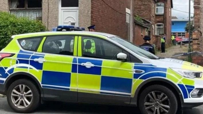 A police car is parked across a cobbled alley where four police officers can be seen with police tape blocking access to the alley.