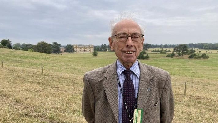 An elderly man in a jacket, shirt, and tie smiles at the camera, wearing a National Trust lanyard, in a field at Croome with the mansion in the background