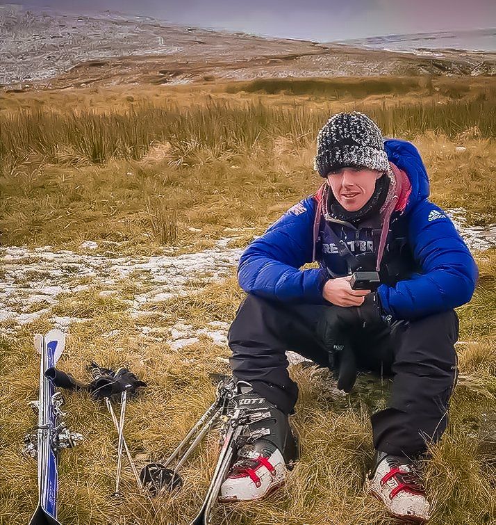 Chris sitting next to his skis in some grass with snow on a mountain in the background