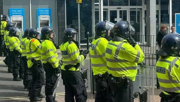 Riot police stand in a line in Stoke-on-Trent, wearing protective blue helmets and fluorescent police jackets, while holding transparent shields