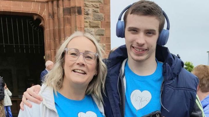 A smiling Caroline Campbell is embraced by her teenage son Oscar as they both stand outside the Guildhall in Derry. They are both wearing blue t-shirts, Caroline is wearing a grey hoodie while Oscar, who is also smiling, is wearing a navy jacket and headphones on his head