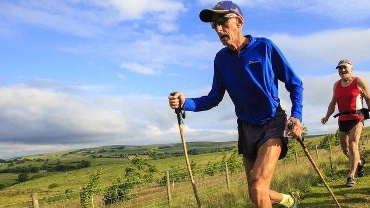 Joss Naylor running on a countryside path, holding two sticks, with a view of fells and another runner behind him.