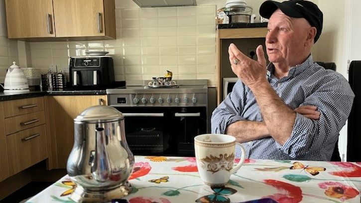 Billy Welch, wearing a baseball cap, sits at a table gesticulating with cups of tea and a teapot on the table