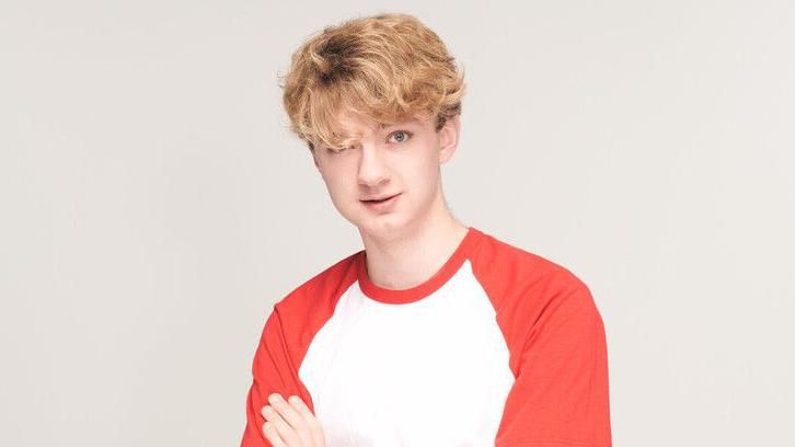A young man with blond curly hair wears a red and white t-shirt and folds his arms