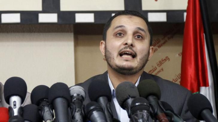 Interior ministry spokesman for the Hamas-run government, Ihab al-Ghussein speaking to journalists in front of a row of microphones. He is seated in front of a Palestine flag.