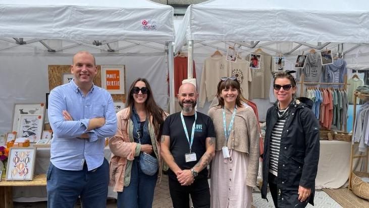 Five people, stood in front of a clothing and art stall atNewquay Market. They are looking at the camera and smiling.