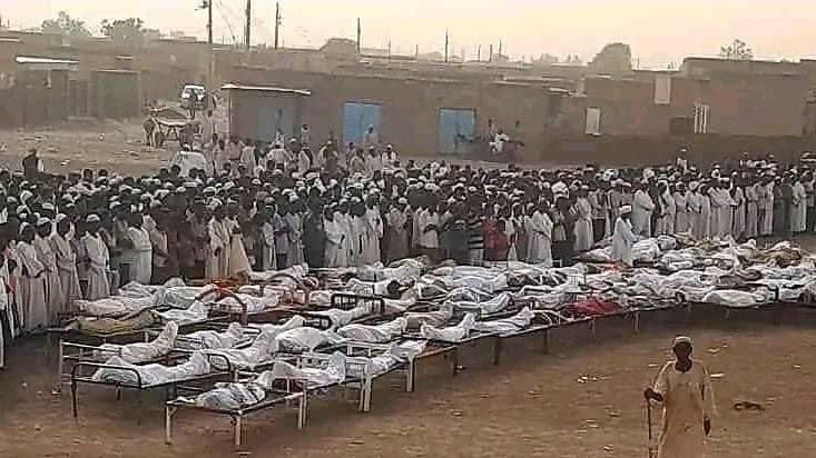 A large group of mourners stand in an open field beside a number of bodies wrapped in cloth.