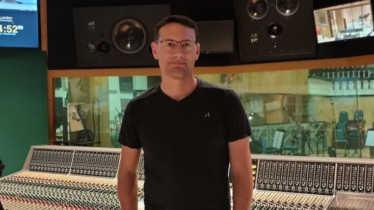 Nick Newton is wearing a black t-shirt and brown trousers. He is standing in front of a large sound mixing desk in a music studio.