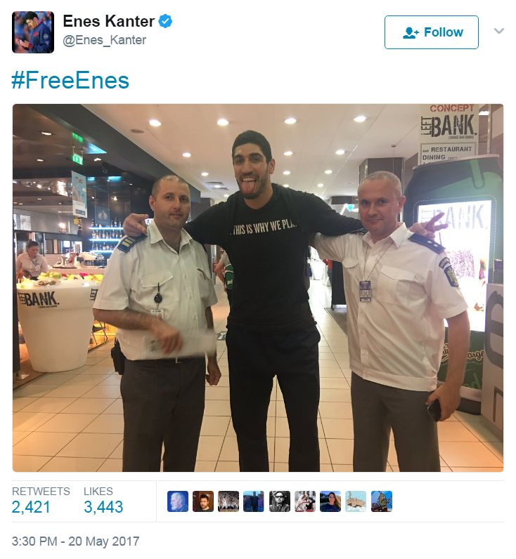 Screenshot from the Twitter account @Enes_Kanter, showing the athlete with an arm around two airport police. The caption reads: #FreeEnes
