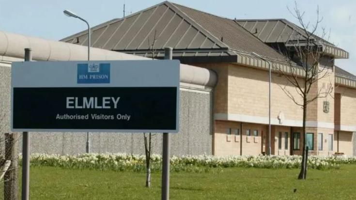 A large stone prison building set in green lawns with a sign reading HMP Elmley outside