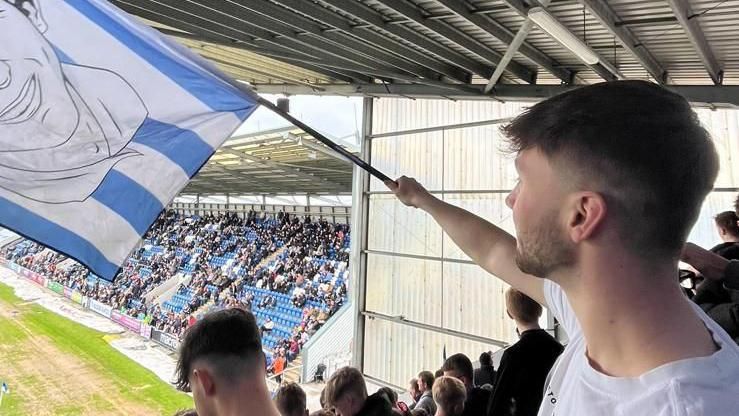 Ian Hart waves a flag in the stands at Colchester United's JobServe Community Stadium