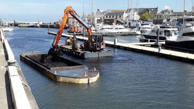 Silt is dredged from Peel marina with a digger on a barge lifting it onto a another barge for transportation. 