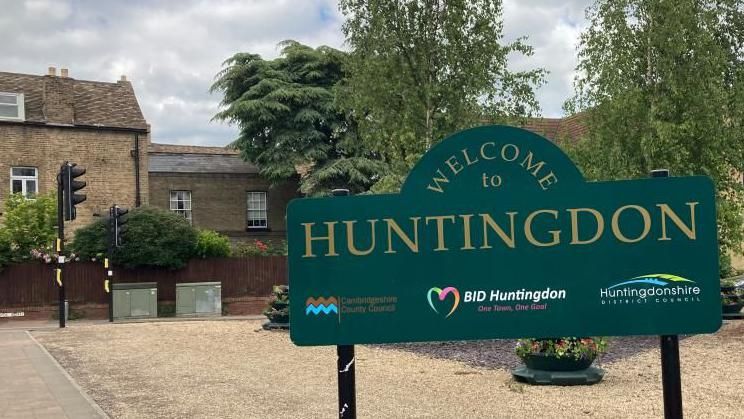 A green sign reading "Welcome to Huntingdon" with a gravel car park behind it and houses