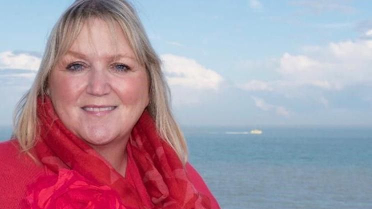 Karen Constantine wearing a red scarf and standing by the sea