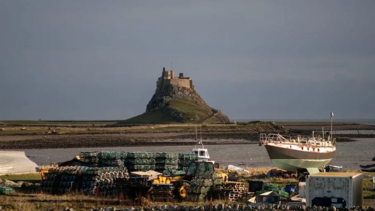 A castle on top of a green hill surrounded by the sea. Boats and fishing equipment can be seen in the foreground.