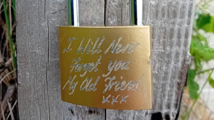 A padlock attached to a wooden post stating "I will never forget you my old friend xxx"