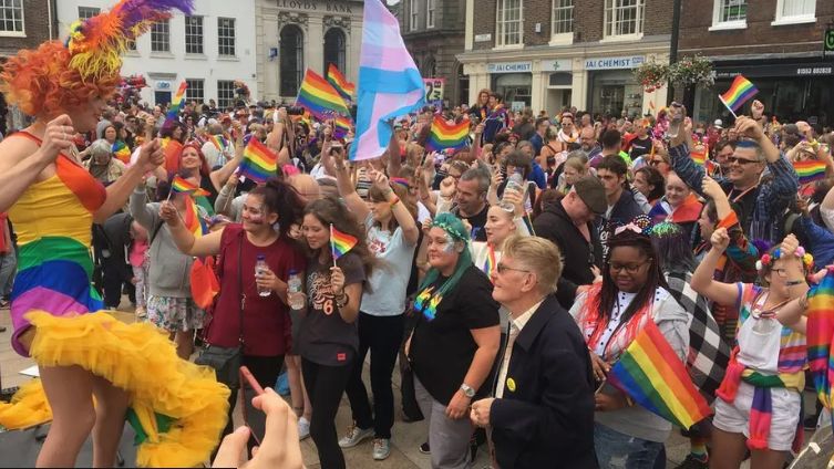 A crowd of people waving rainbow flags watching Titania Trust in a rainbow dress in Tuesday Market Place