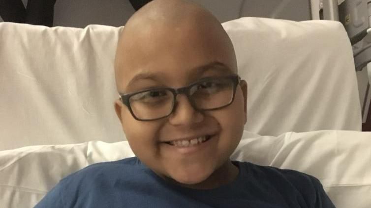 A little boy with no hair and dark rimmed glasses wears a navy t-shirt and smiles broadly at the camera from a hospital bed
