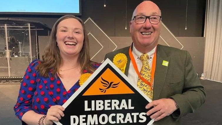 Smiling Kerrie Bradburn wearing a blue and pink spotted dress and a yellow Lib Dem party rosette. She is holding a sign saying Liberal Democrats with her father. He also has a yellow rosette and a yellow tie with winged birds on in black and is smiling. 