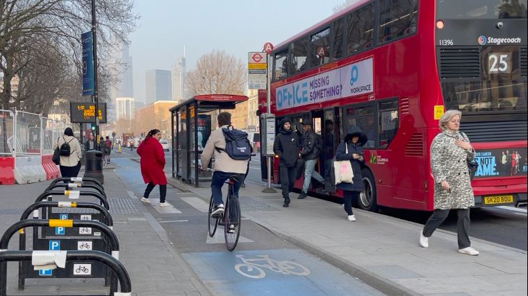 A pedestrian crosses a cycle lane to reach a floating bus stop, she is wearing a red coat and is stepping back slightly as a cyclist approaches in the cycle lane. On the right is a bus stop with a red London bus with passengers coming off.