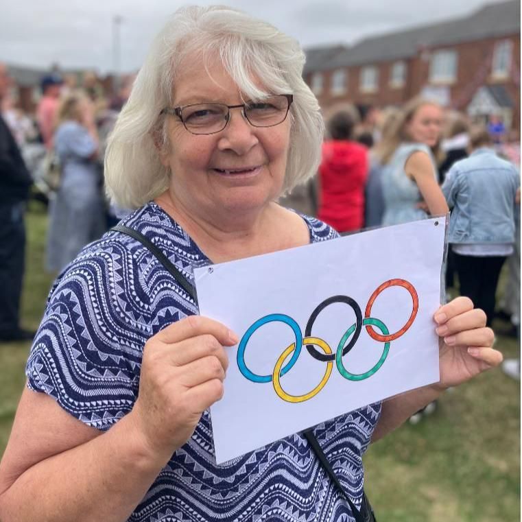 A woman wearing a blue patterned shirt holding a piece of paper with Olympic rings on