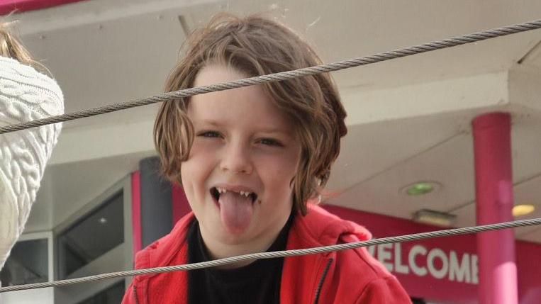 Lennix pictured sticking his tongue out at the camera, wearing a black tshirt and a bright red hoodie. 