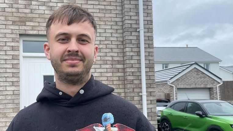 Connor Davidson standing in front of his brick house. he wears a Motley Crue Hoody and has brown hair and stubble. 