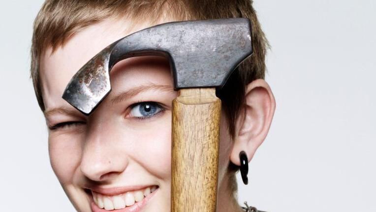 A young woman smiles whilst holding up a wooden tool
