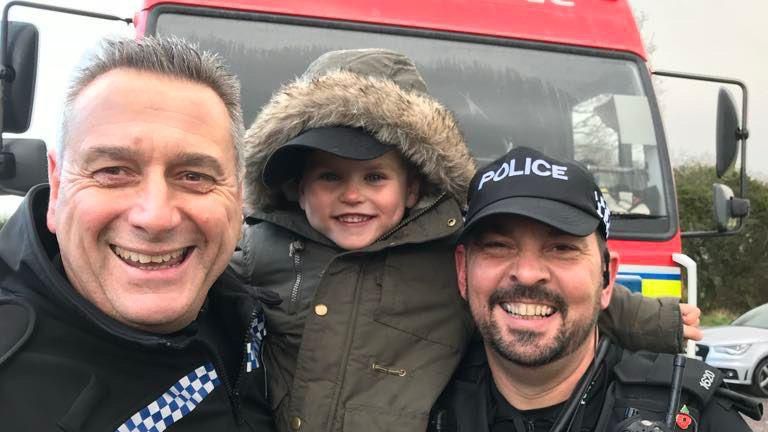 A five-year-old Jayden being held up by two policeman, all smiling and looking into the camera