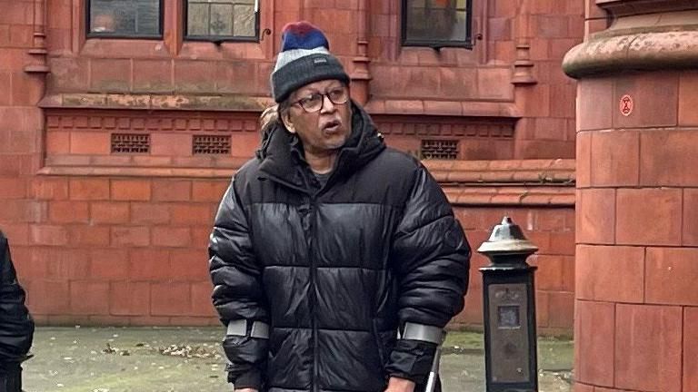 A man in a black puffer jacket and woolly hat and wearing glasses walking with crutches outside Birmingham Crown Court