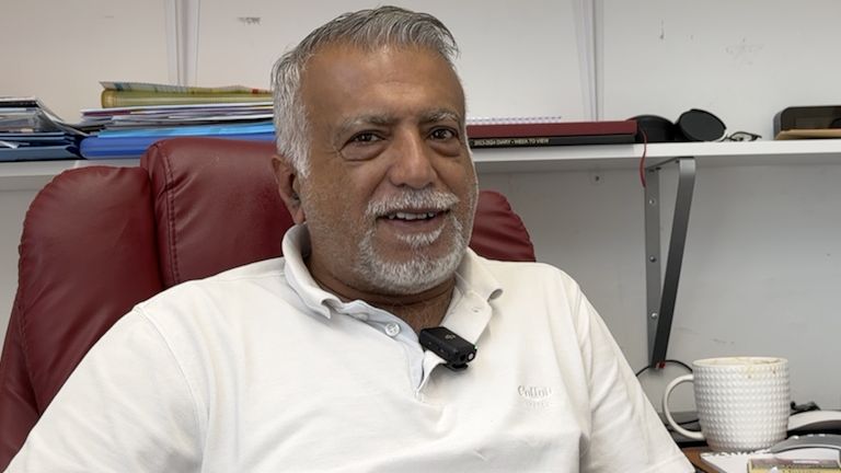 Shop owner Bipin Chawla, who is a middle aged Indian man, with grey hair and a grey short beard,  sat in his shop office in a red leather computer chair, wearing a white polo shirt, with a white coffee mug showing on his desk next to him