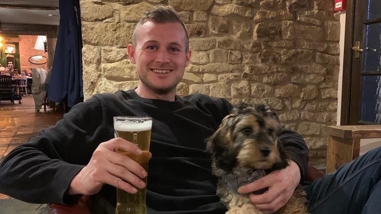 Tom Sturgess with short brown hair with a pint in one hand and a dog in the other
