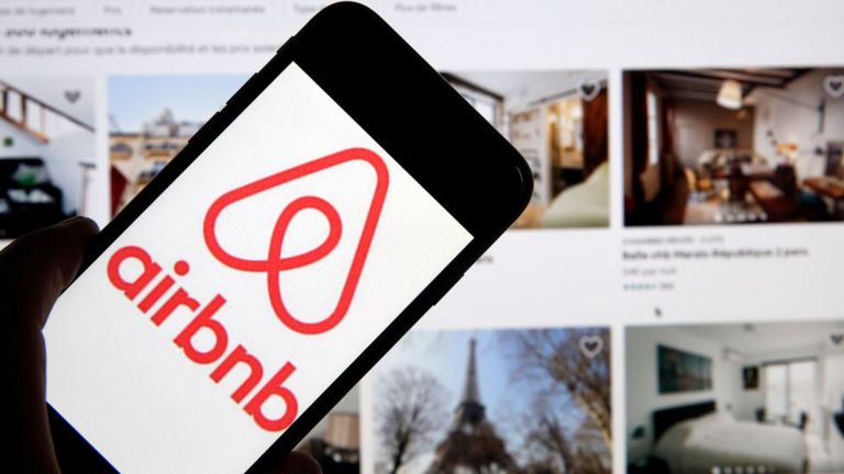 A man holding a phone displaying the Airbnb logo, in front of the firm's website