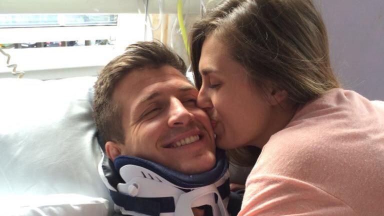 Ed Jackson lying in a hospital bed wearing a neck brace. His partner, Lois, is kissing his cheek. 