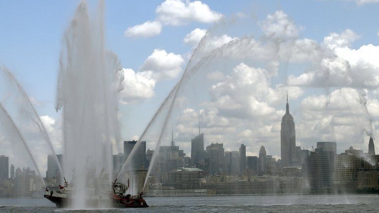 The New York City skyline and the Empire State Building are visible under the spray from a fire boat