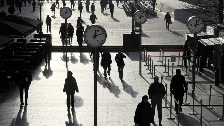 Workers walk past clocks at Canary Wharf