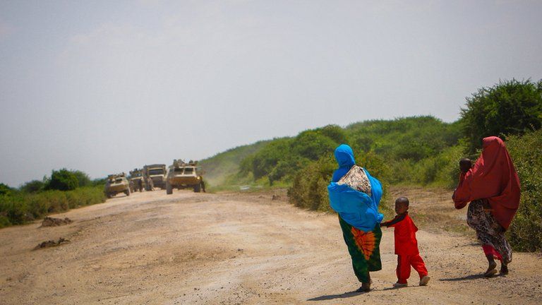 Somali women and their children walk along a road with security forces vehicles in the background