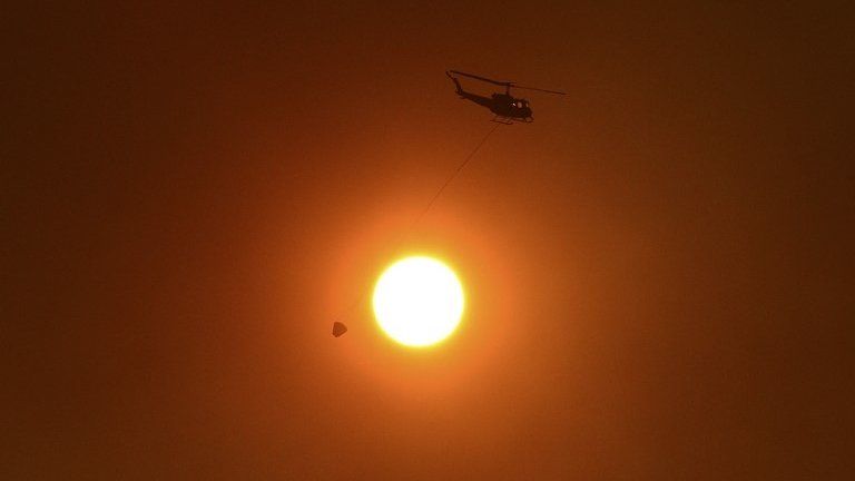 A helicopter with a bucket used for carrying water for dropping onto bushfires, flies past the sun as it is obscured by smoke in western Sydney 20 October 2013