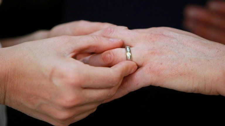 A close-up of hands as one person puts a wedding ring on the finger of another