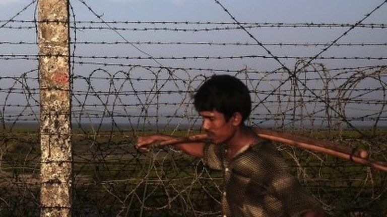 A Rohingya worker carries wood alongside a barbed wire fence.