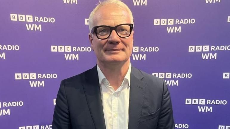 Richard Parker in black jacket, white shirt and glasses, standing against a purple background with the BBC Radio WM logo across it