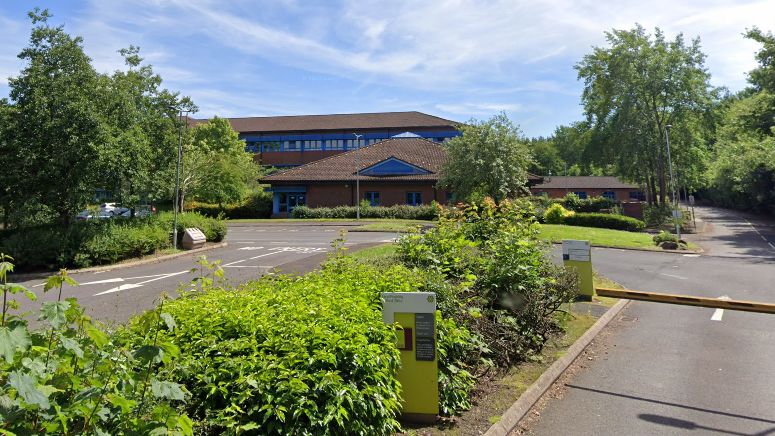 A brown brick building with glass windows is in the background, behind green trees and bushes. There is a yellow traffic barrier leading into a car park, and a yellow sign in front of it. The sky is blue with some clouds.