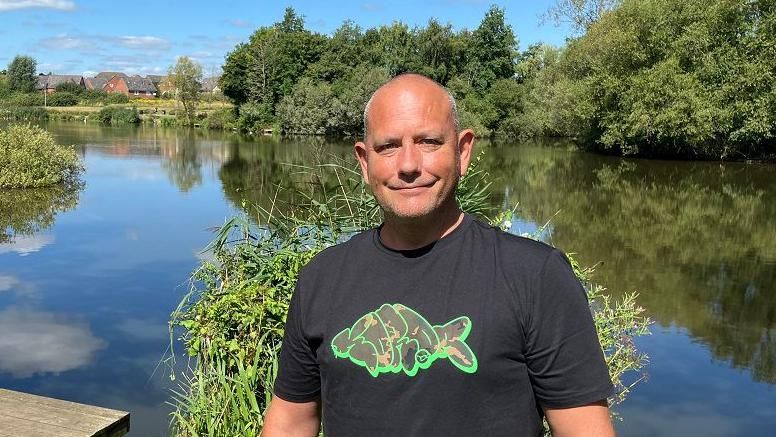 Michael Bromley stands in front of a lake on a sunny day. He is wearing a black t-shirt with a motif of a fish and looking into the camera.