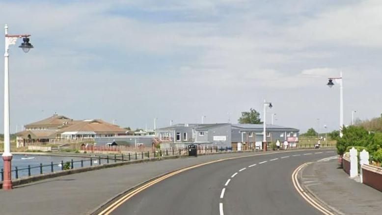 The Promenade in Southport - a road winds to the right - lined with double yellow lines. On the left is water and some grey buildings. on the right is gates into a residential home with green bushes. 