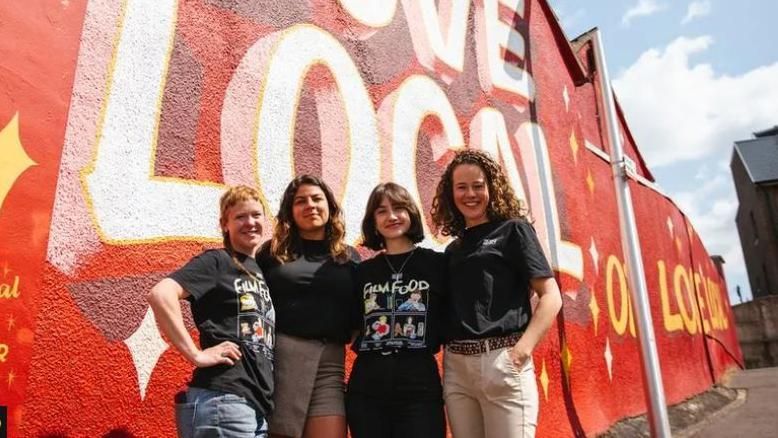 Four women in black t shirts stand in front of a house painted with a red mural