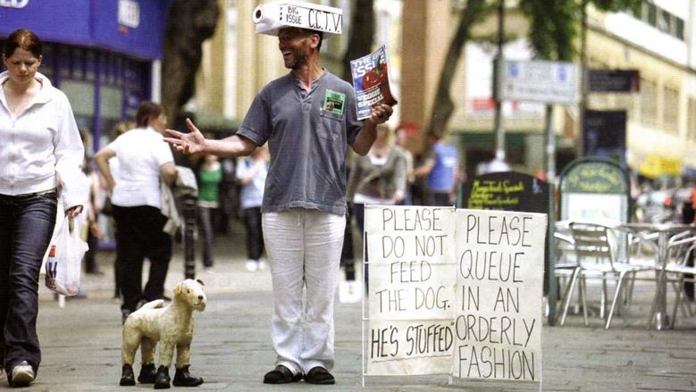 Mr Walker selling the Big Issue next to a stuffed dog, a board with signs on and wearing a mock CCTV camera on his head