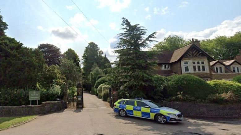 Entrance and driveway to cemetery with trees either side and a police car in the foreground